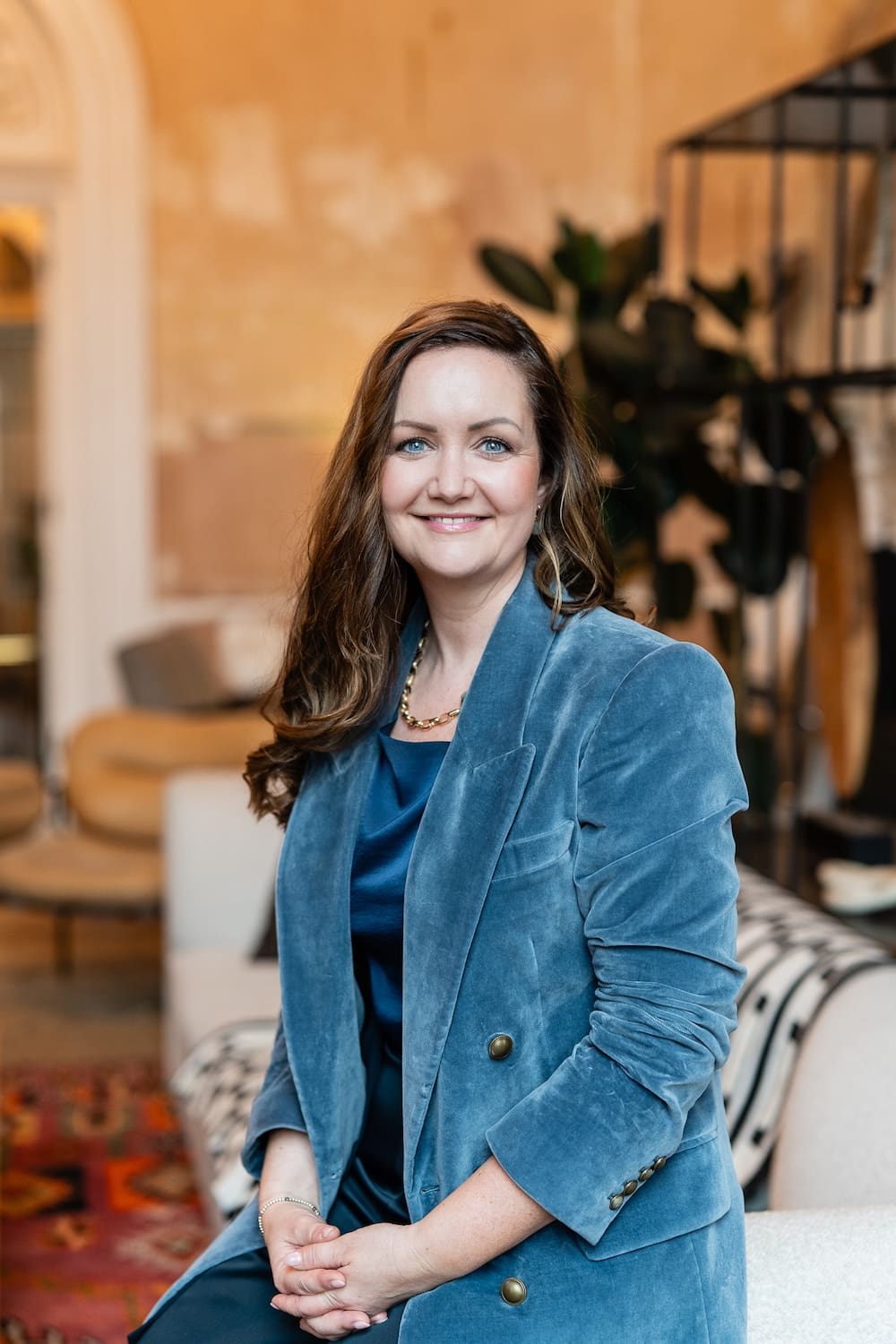 woman in blue shirt and blue blazer sitting on a beige sofa and smiling